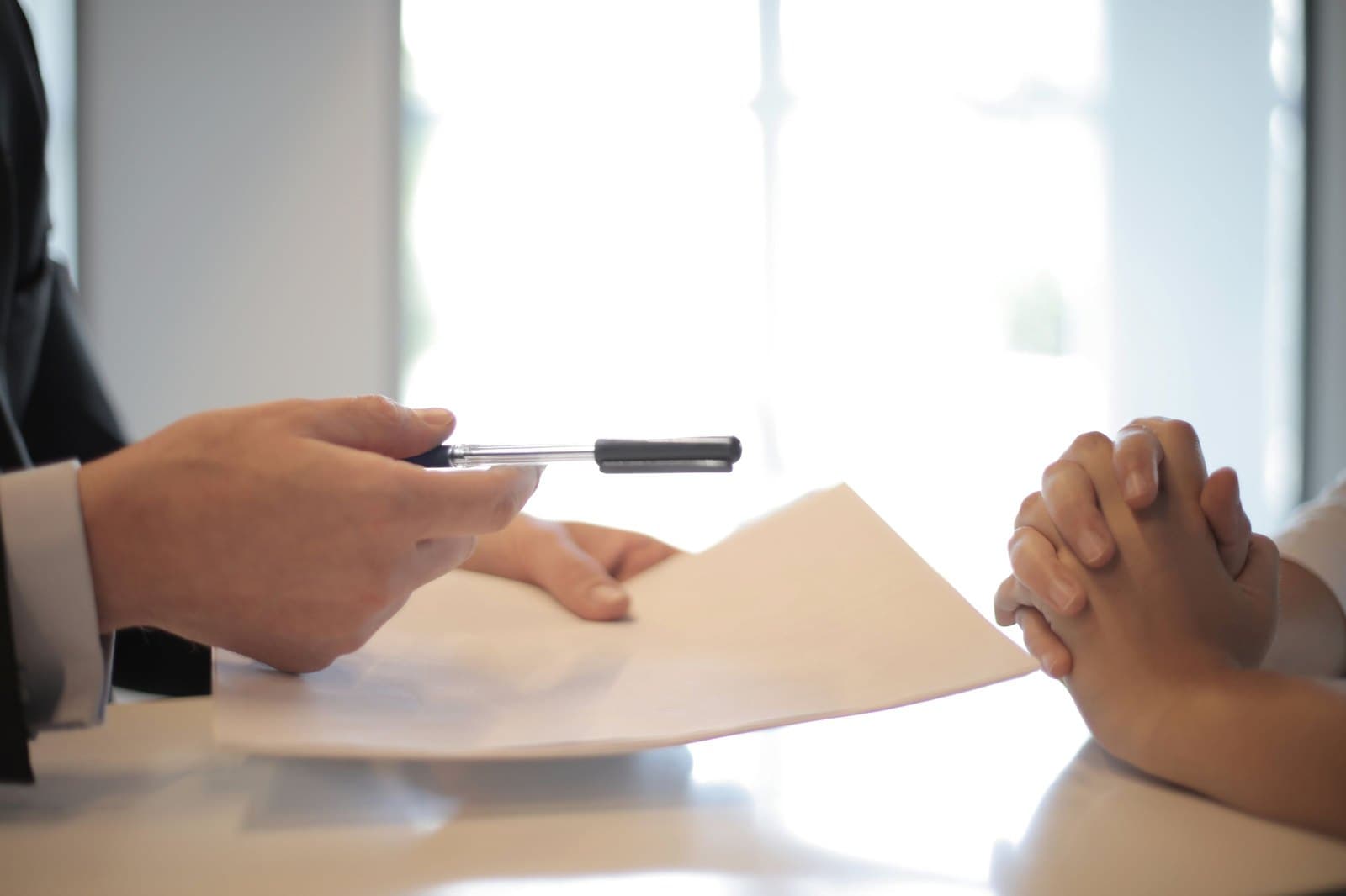 A person in a suit offering a pen and a blank page across a desk.