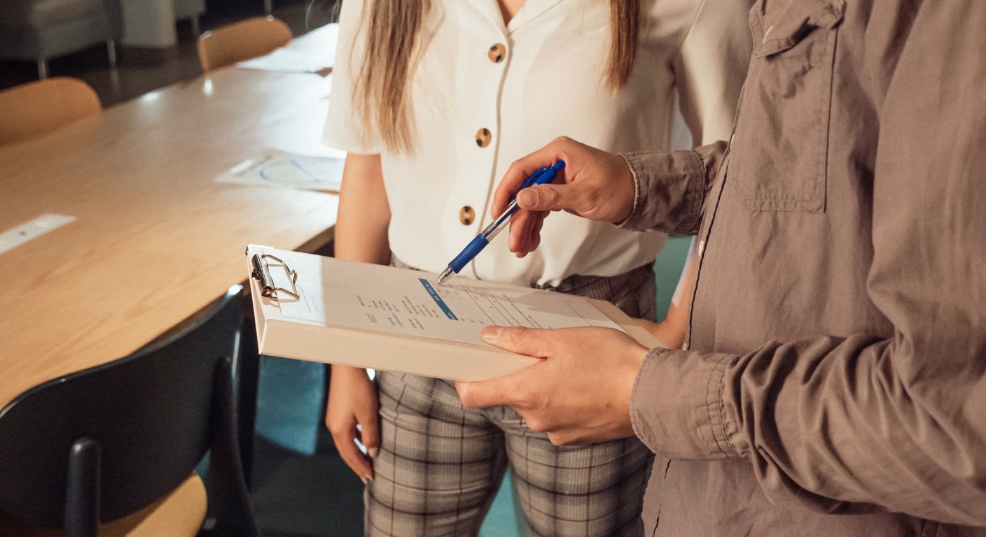 Two people in an office review a multi-page document held open in a blue binder; one person points to a line on the page.