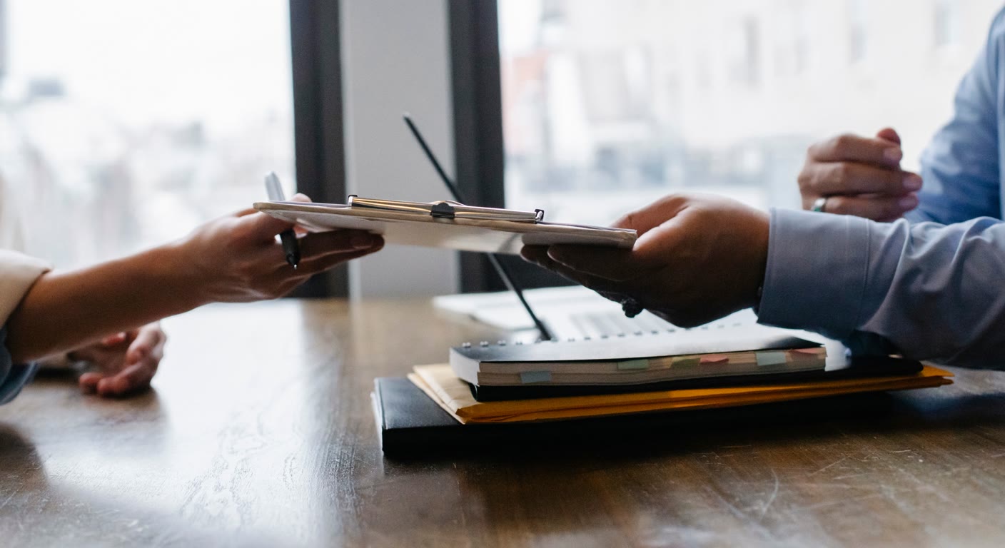 Two people exchanging a clipboard of papers over a desk next to a tabbed notebook