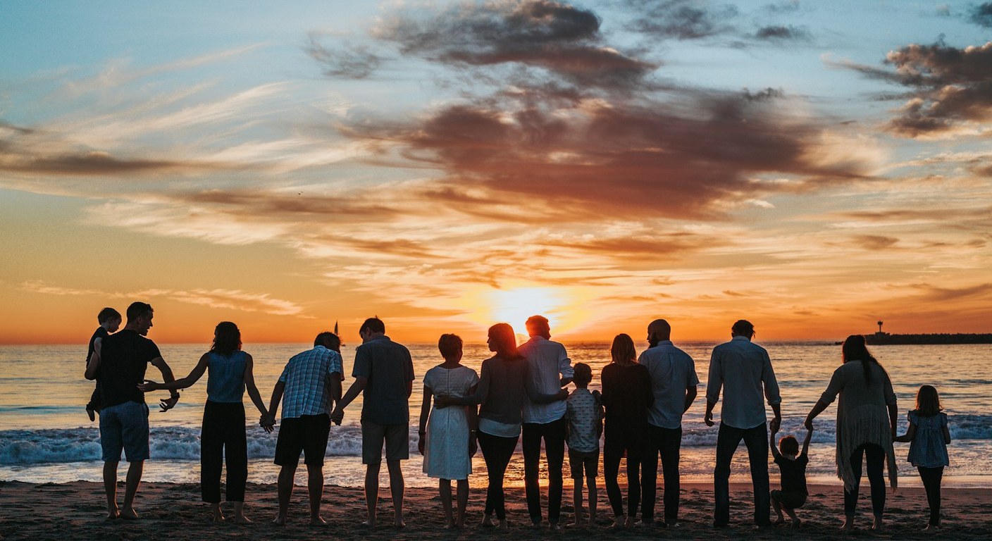 Multi-generational family holding hands as silhouettes against a bright sunset on a beach.