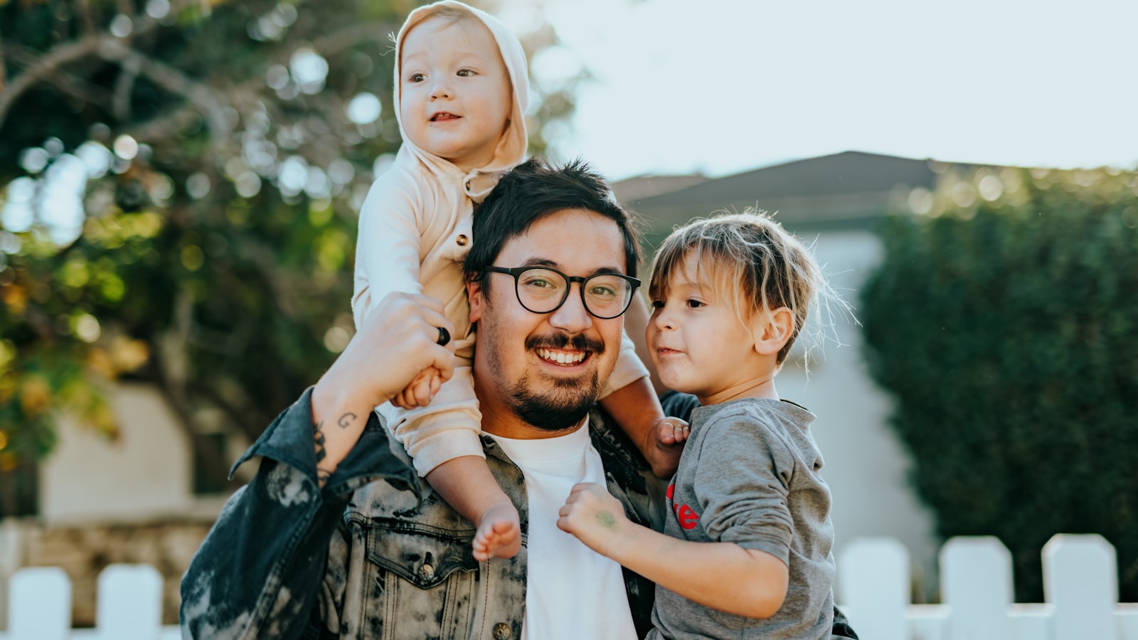 A smiling parent outdoors holds two young children near a white fence.