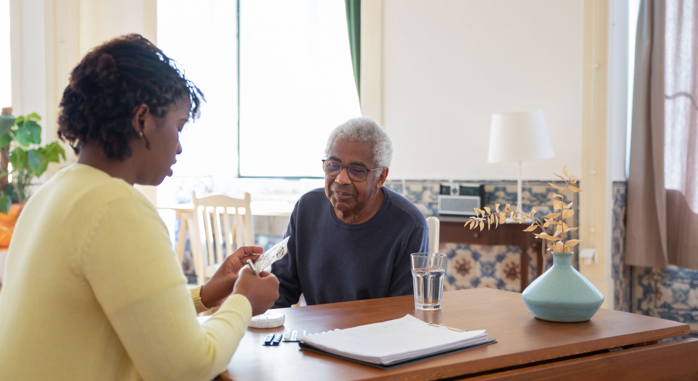 Younger caregiver in a yellow sweater preparing blister-pack medication across a wooden table from an older man in glasses in a sunlit home room, with a glass of water and pill organizer on the table.