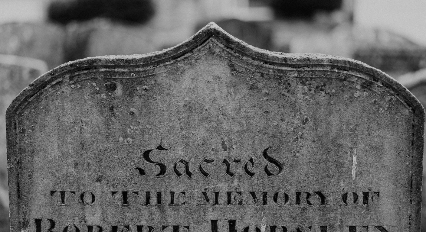 Close-up of a weathered private stone headstone with the carved words Sacred to the memory of, soft background of other cemetery markers.