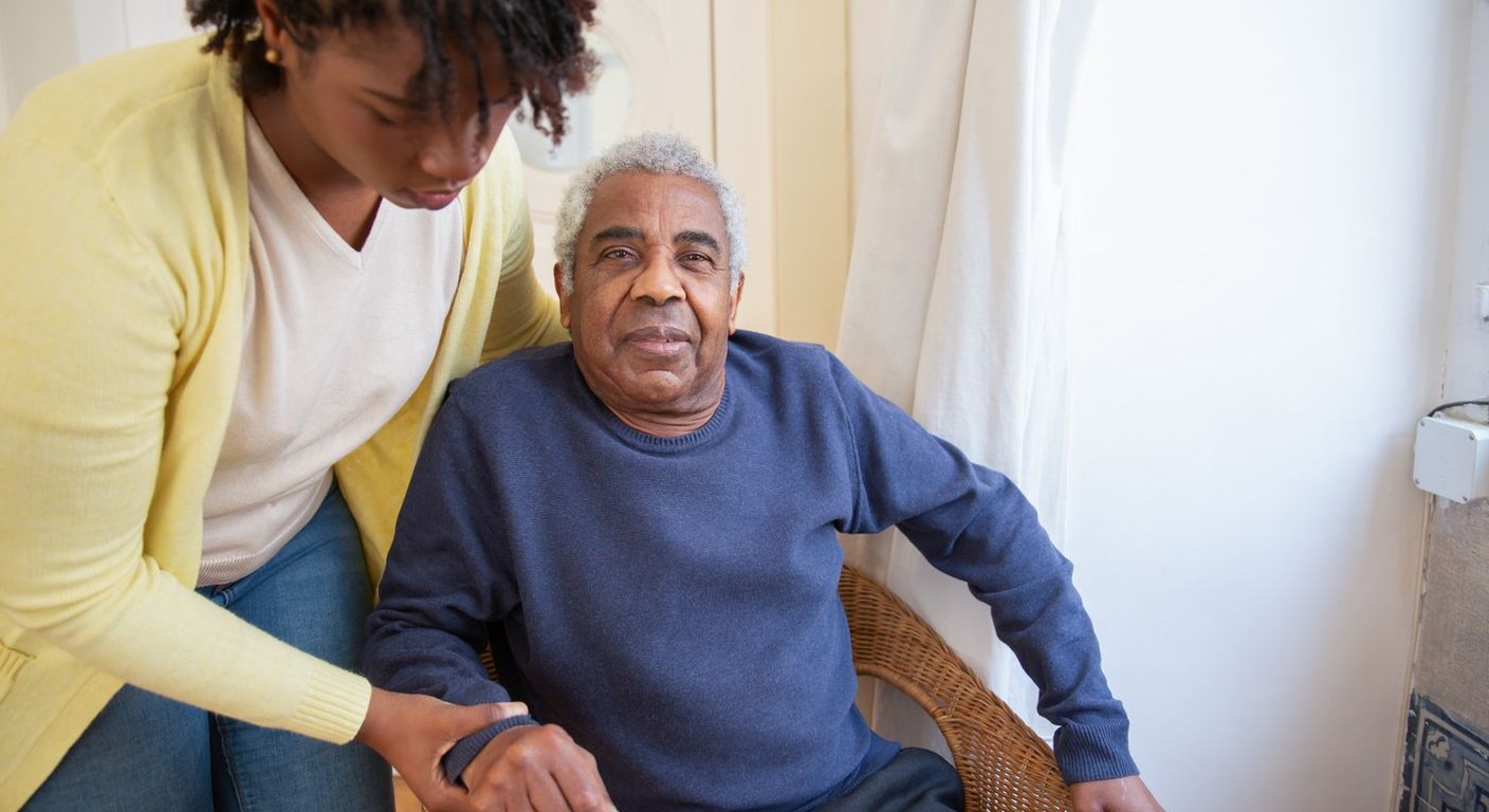 Younger caregiver in a pale yellow cardigan gently supporting an older man’s wrist as he sits in a wicker chair in a bright home room.